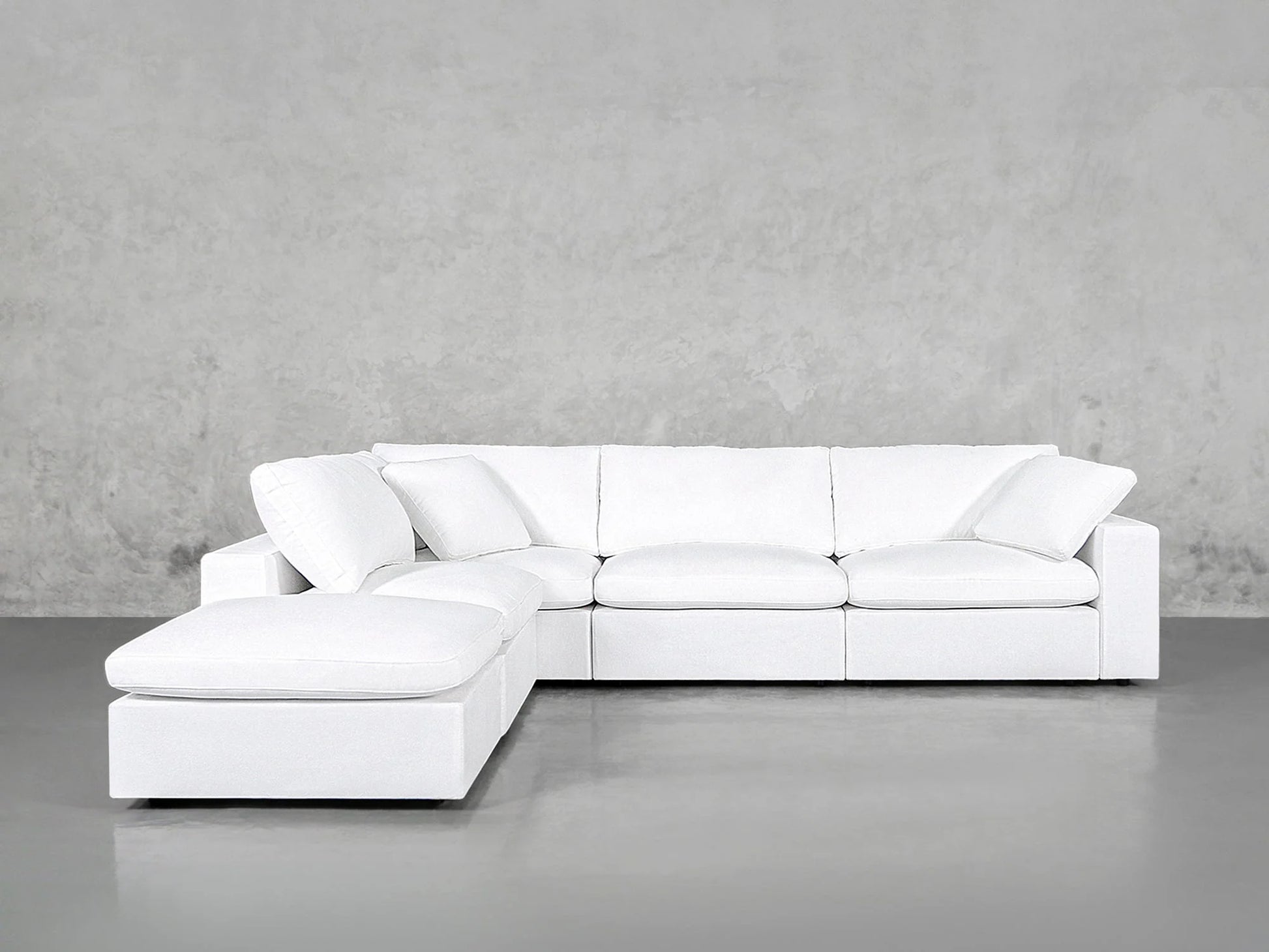 A contemporary white 5-Seat Modular Corner Lounger Sectional with cushioned seats and backrests is positioned against a plain gray concrete wall. The smooth gray flooring enhances the minimalist style of the room.