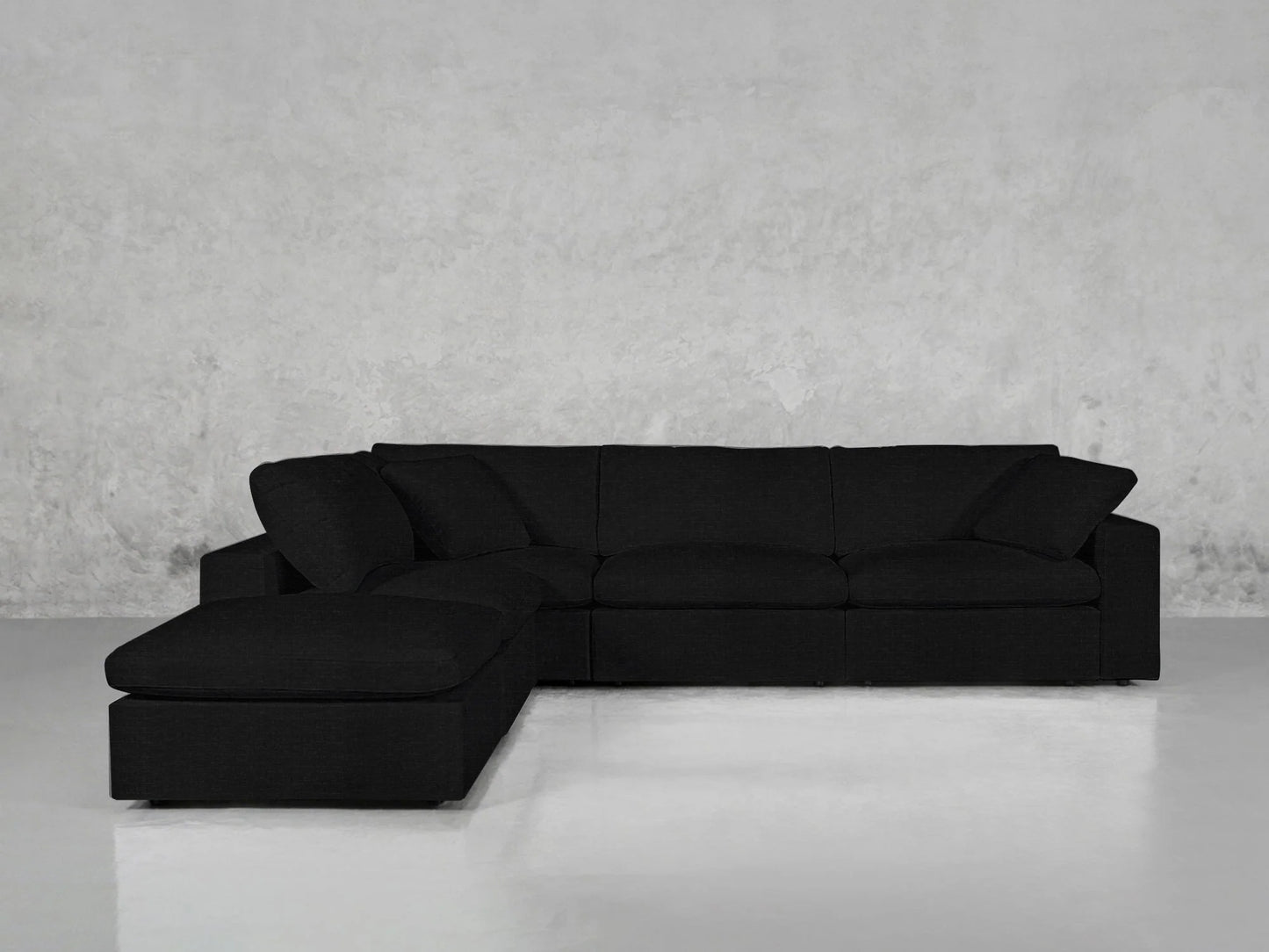 The 5-Seat Modular Corner Lounger Sectional in sleek black is positioned against a simple gray wall and a glossy light gray floor, featuring a chaise lounge on the left.