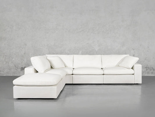 A 5-Seat Modular Corner Lounger Sectional, characterized by its modern style and white hue, sits in a minimalist room with light gray walls and a polished concrete floor. The sectional consists of three main sections and includes a chaise lounge on the left side.