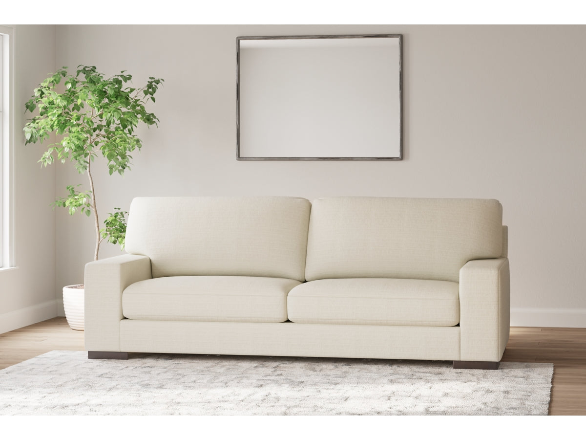 A minimalist living room showcases the Maggie Sofa in white adorned with two cushions, set on a light gray area rug. To the left is a potted plant, while an empty rectangular mirror hangs above the sofa on the wall. Natural light streams in from a window.