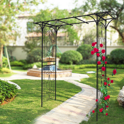 The Metal Garden Arch Climbing Plants Support, a black rose arch, features intricate designs perfect for climbing red roses. It elegantly arches over a curved stone path leading to a circular water fountain surrounded by lush green grass and bushes.