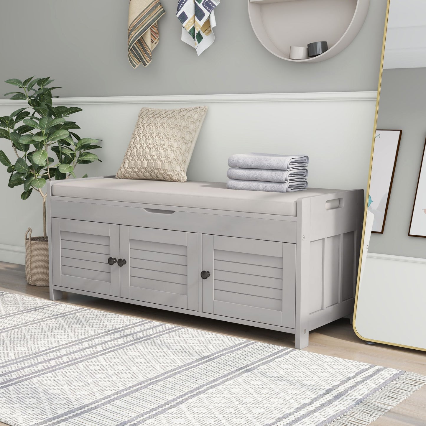 A white storage bench with three shutter-shaped doors and a removable cushion sits against a light green wall, topped with folded towels and a pillow. Nearby are a potted plant, round mirror, wall hooks with hanging items, and a large gold-framed mirror.