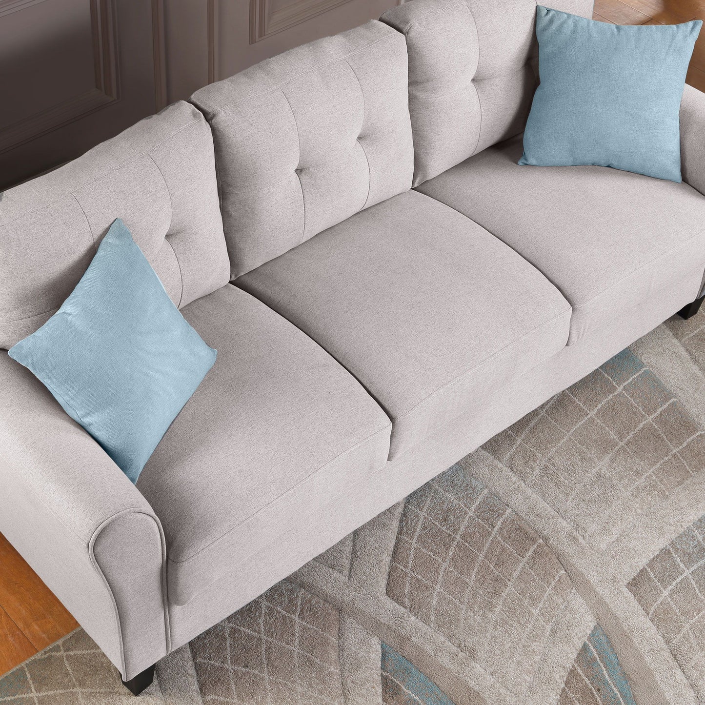 The Modern Living Room Loveseat, a light gray sofa with tufted cushions and armrests, features two light blue throw pillows and is placed on a patterned area rug with beige, gray, and blue shades atop a hardwood floor.