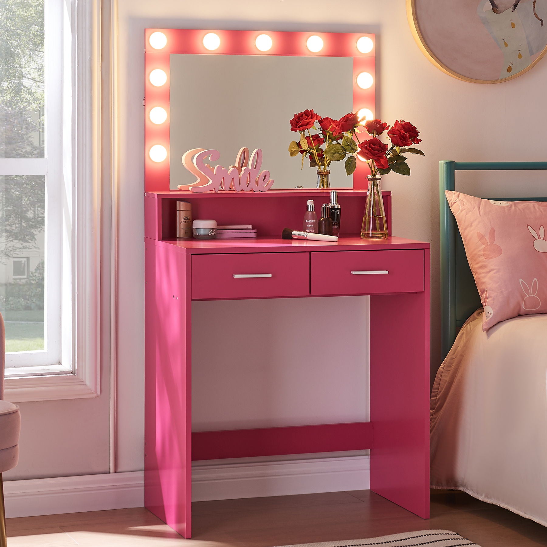 A Vanity Desk with Mirror and Lights, featuring a large drawer and two-level storage, is decorated with red roses, a "Smile" sign, and makeup items. Positioned by a window next to a pink bed, it adds brightness and cheer to the bedroom corner.