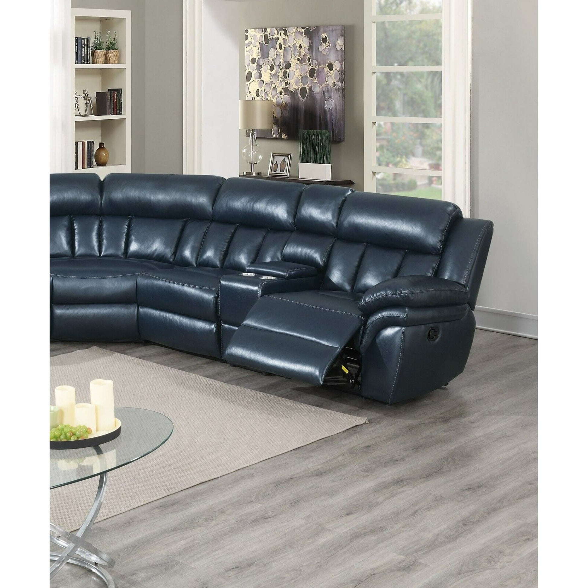 A modern living room features the Luxurious Manual Motion Sectional Sofa in a large, dark blue leather design. One side of the sofa is reclined. The space includes a round glass coffee table adorned with candles, accompanied by a bookcase and wall art, while large windows provide a view of the outdoors. The room boasts light wood flooring.