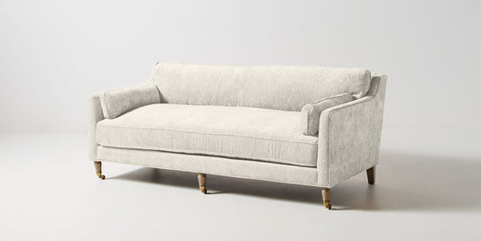 The Leonelle Sofa, a light gray upholstered piece featuring wooden legs and brass casters, is set against a plain white background. It boasts a minimalist design with plush cushions and comfortable armrests.