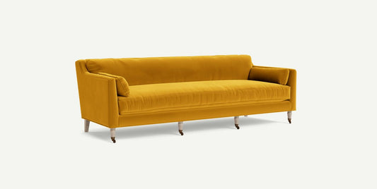 A Leonelle Sofa in mustard yellow velvet, featuring three cushions and wooden legs with casters, set against a plain white background.