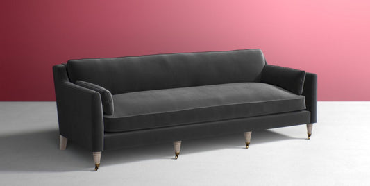 The Leonelle Sofa, featuring a minimalist design with clean lines and plush cushions, is placed against a stylish matte pink wall on a light gray floor. This modern gray sofa with wooden legs offers a comfortable and elegant seating option.