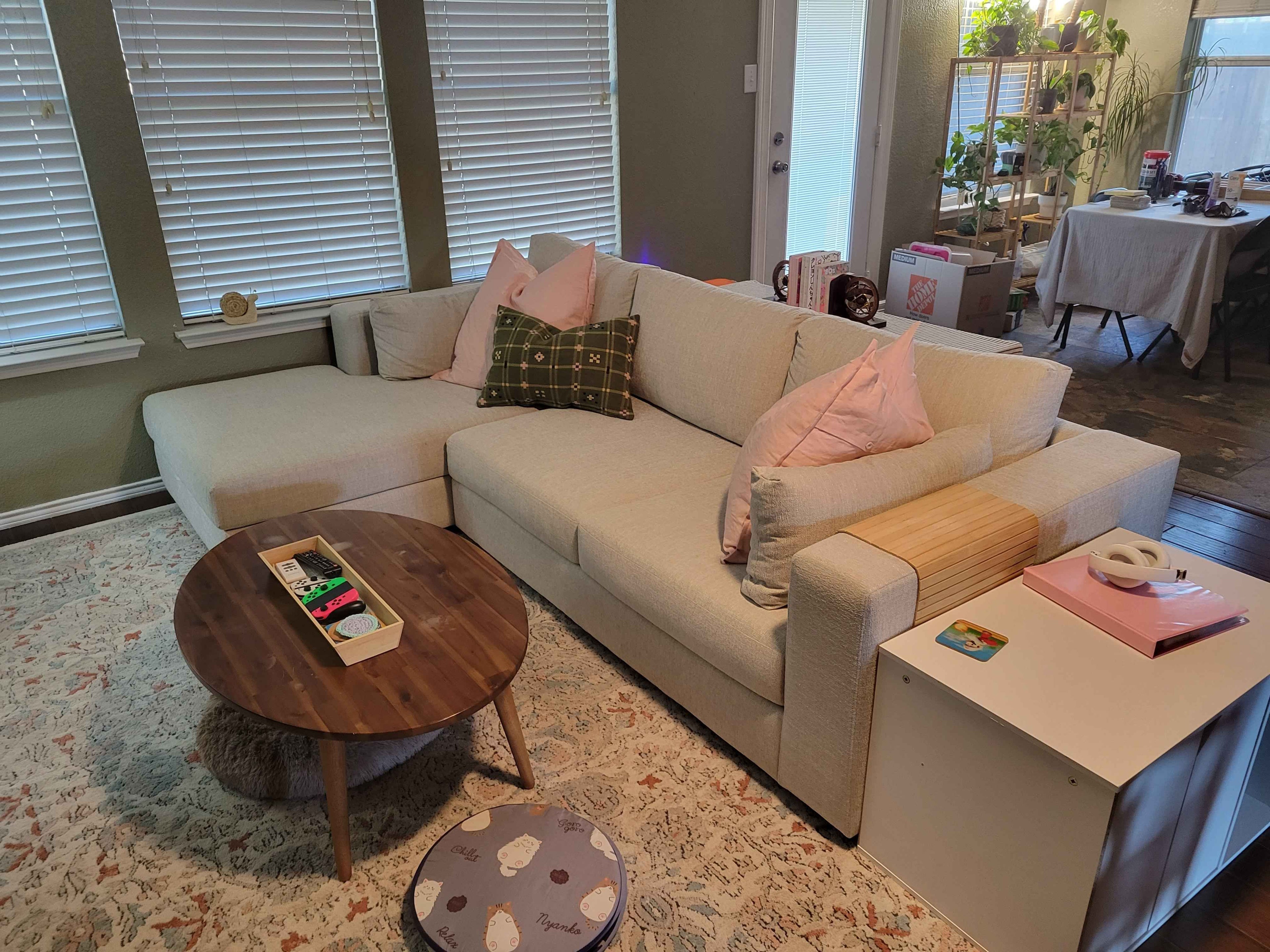 A cozy living room with a light beige sectional sofa adorned with pink and plaid pillows. A round wooden coffee table holds small items. A soft rug covers the floor, and the room is brightened by large windows and greenery in the background.