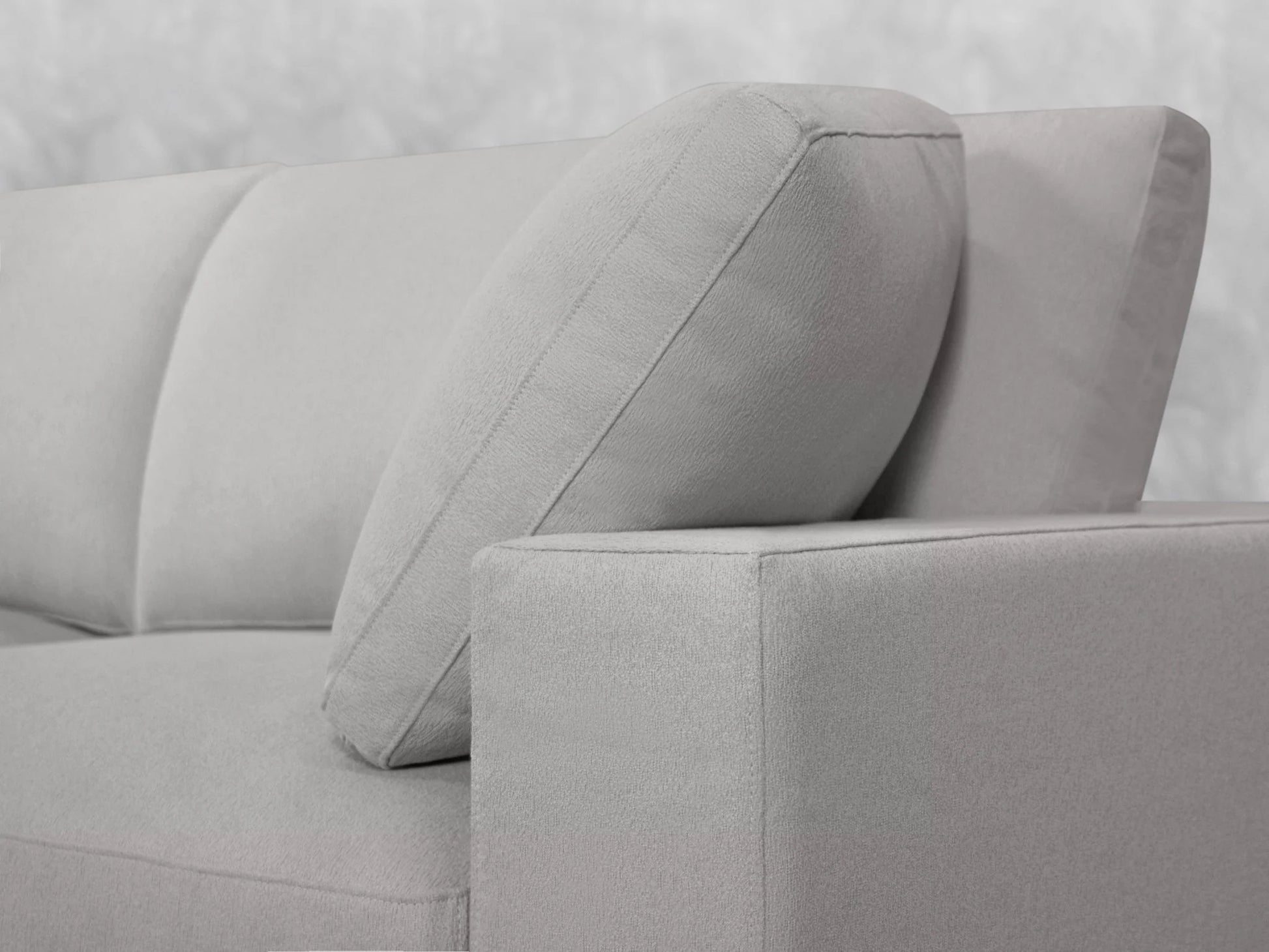 Close-up of a 4-Seat Modular Chaise Sectional with Ottoman in a modern, light gray fabric. It features clean lines and plush cushions, set against a neutral background. The focus is on the corner section, highlighting the sectional's minimalist design.