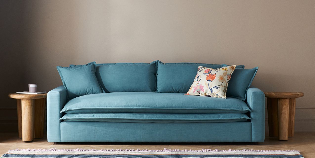 The Meriwether Sofa in teal, complete with multiple cushions including a floral-patterned one, is positioned between two round wooden tables set against a neutral wall. A small object is placed on the table to the left. A fringed rug can be partially seen in the foreground.