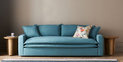 The Meriwether Sofa in teal, complete with multiple cushions including a floral-patterned one, is positioned between two round wooden tables set against a neutral wall. A small object is placed on the table to the left. A fringed rug can be partially seen in the foreground.