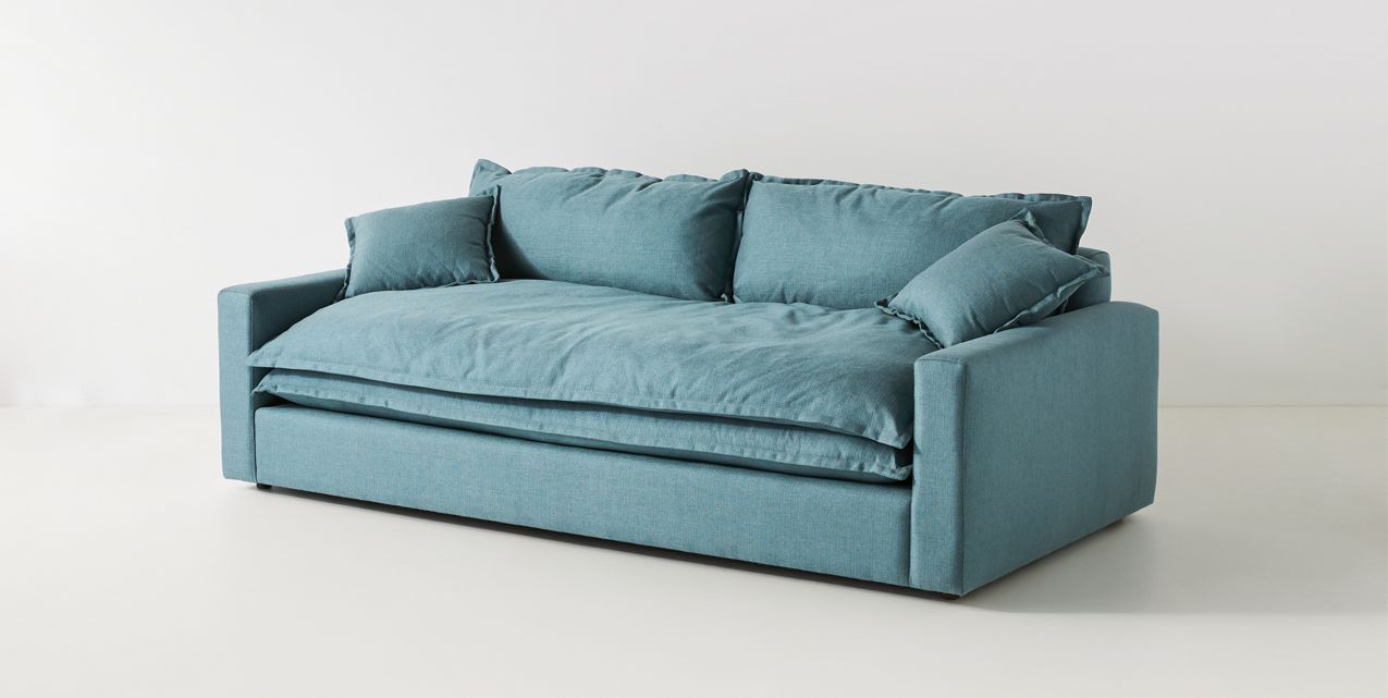 The Meriwether Sofa, with its minimalist design and teal color, is set against a white background. It boasts a large cushioned seat and three matching pillows on the back, providing a cozy and modern appearance.