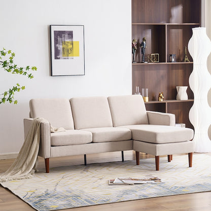 A modern living room featuring the Outopee Modern Beige Linen Sectional paired with an ottoman on a patterned rug. In the background stands a tall wooden shelf adorned with decor, complemented by a unique tall lamp. Above the sofa, a framed abstract art piece is displayed on the white wall.