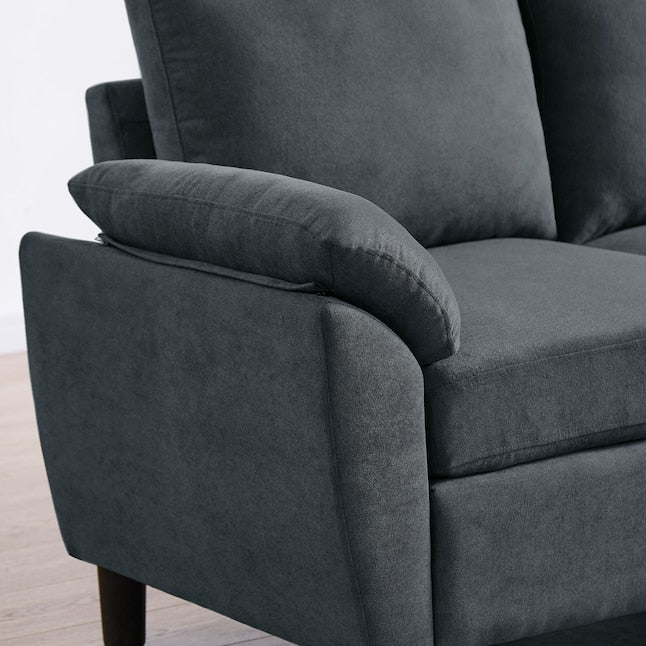 Close-up of the Jarenie Casual Dark Grey Linen Sectional with plush cushions and wooden legs. The image captures the armrest and side profile, showcasing its soft linen fabric texture. The background features a light-colored floor and wall.