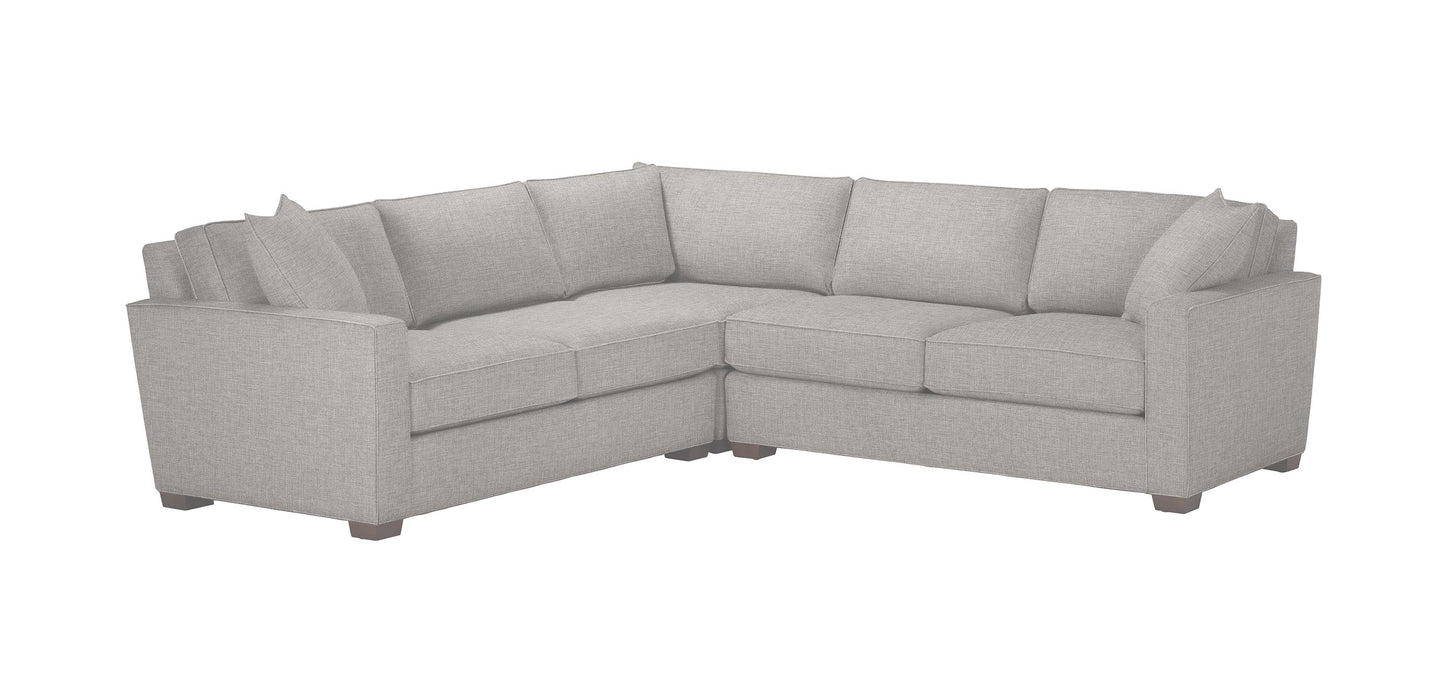 The Spencer Track-Arm 3-Piece Sectional, Quick Ship is a light gray, L-shaped sofa featuring plush cushions and low armrests. Its design showcases visible seams and square wooden legs, offering a modern and cozy aesthetic.