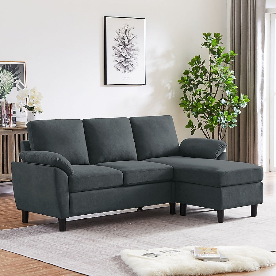 The Jarenie Casual Dark Grey Linen Sectional, featuring a chaise lounge, is placed in a bright living room. It rests on a light rug against a white wall decorated with art and adorned by a potted plant. Nearby are a sideboard and curtains framing the window.