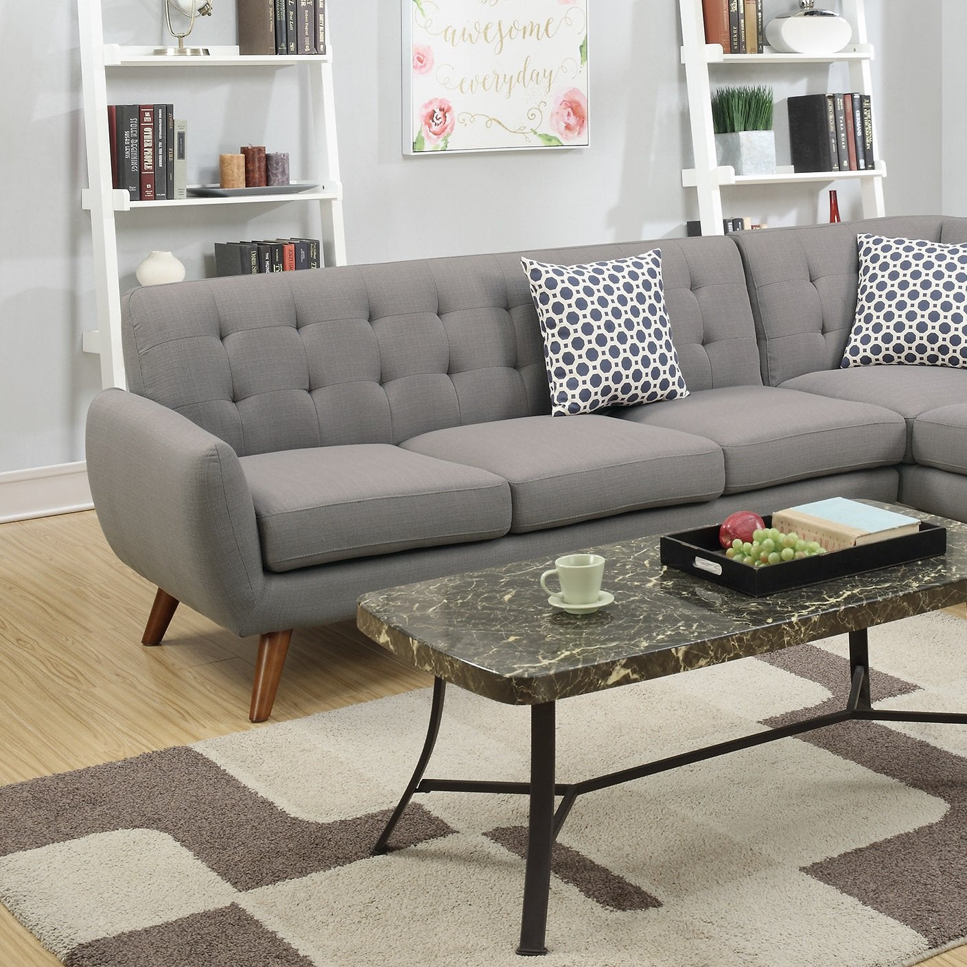 A contemporary living room featuring a Retro Modern Grey Sectional Sofa adorned with patterned pillows. In front, a marble coffee table displays a tray with a green apple and grapes, alongside a cup, while bookshelves and wall art enhance the background decor.