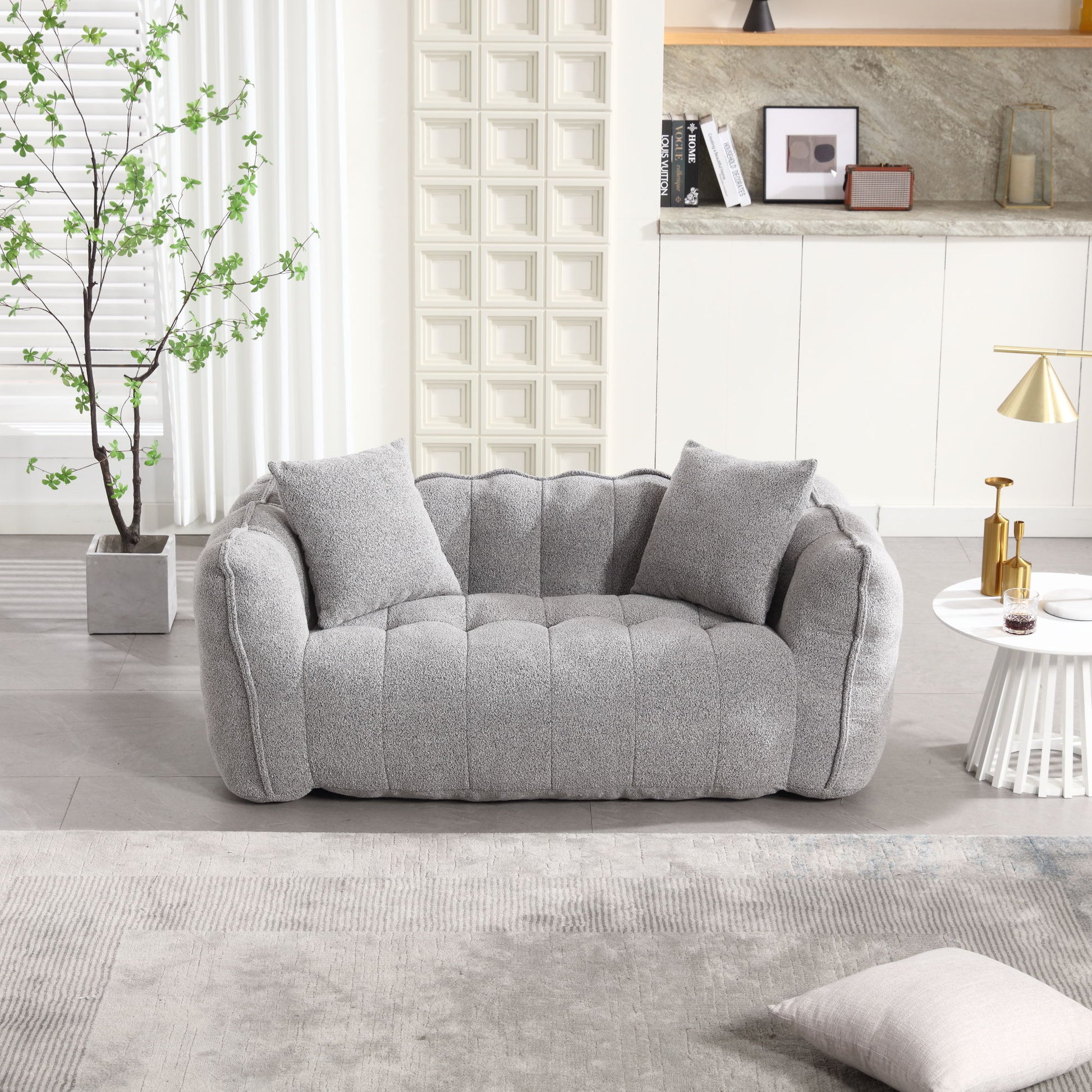 A modern living room features a plush gray sofa, two matching pillows, a round white side table with decor, and a potted plant. Light walls, geometric paneling, and built-in shelving filled with books and decor are in the background. A soft beanbag chair for two adds comfort and style.