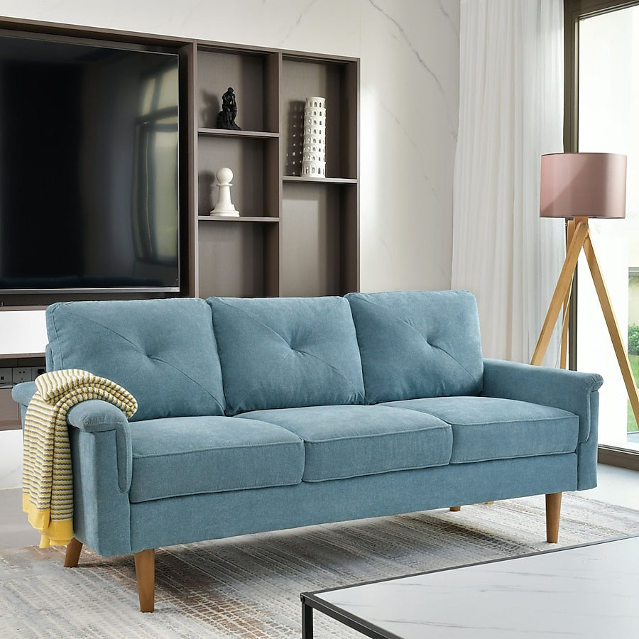 A modern living room features the Ovios 71.65-in Modern Brown Faux Leather 3-seater Sofa with wooden legs. A yellow-striped blanket is draped over the armrest. Behind the sofa is a large TV and an open shelf with decorative items. A floor lamp stands beside a window with white curtains.