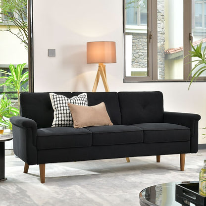 A contemporary living room showcases a modern brown faux leather 3-seater sofa from Ovios, accompanied by three cushions—one with a geometric black-and-white pattern and another in plain beige. Complementing the space is a wooden tripod lamp, positioned nearby, while large windows frame a view of a stone building and flood the room with natural light.