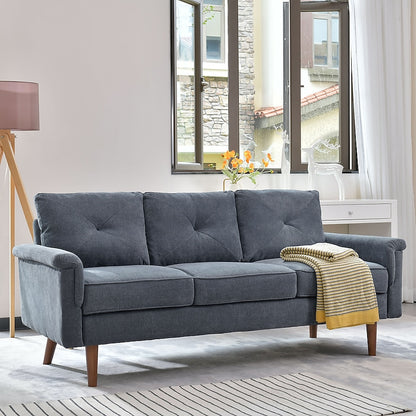 A stylish brown faux leather 3-seater sofa from the Ovios collection is placed in a well-lit living room. A folded yellow and gray striped blanket rests on one arm. Large windows and a white dresser with a vase of flowers on top can be seen in the background, alongside a nearby floor lamp.