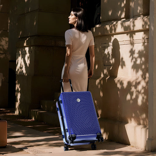 A woman in a white dress strolls along a sunny street, pulling an expandable 3-piece hardshell suitcase with wheels. Tree leaf shadows create patterns on the nearby wall.