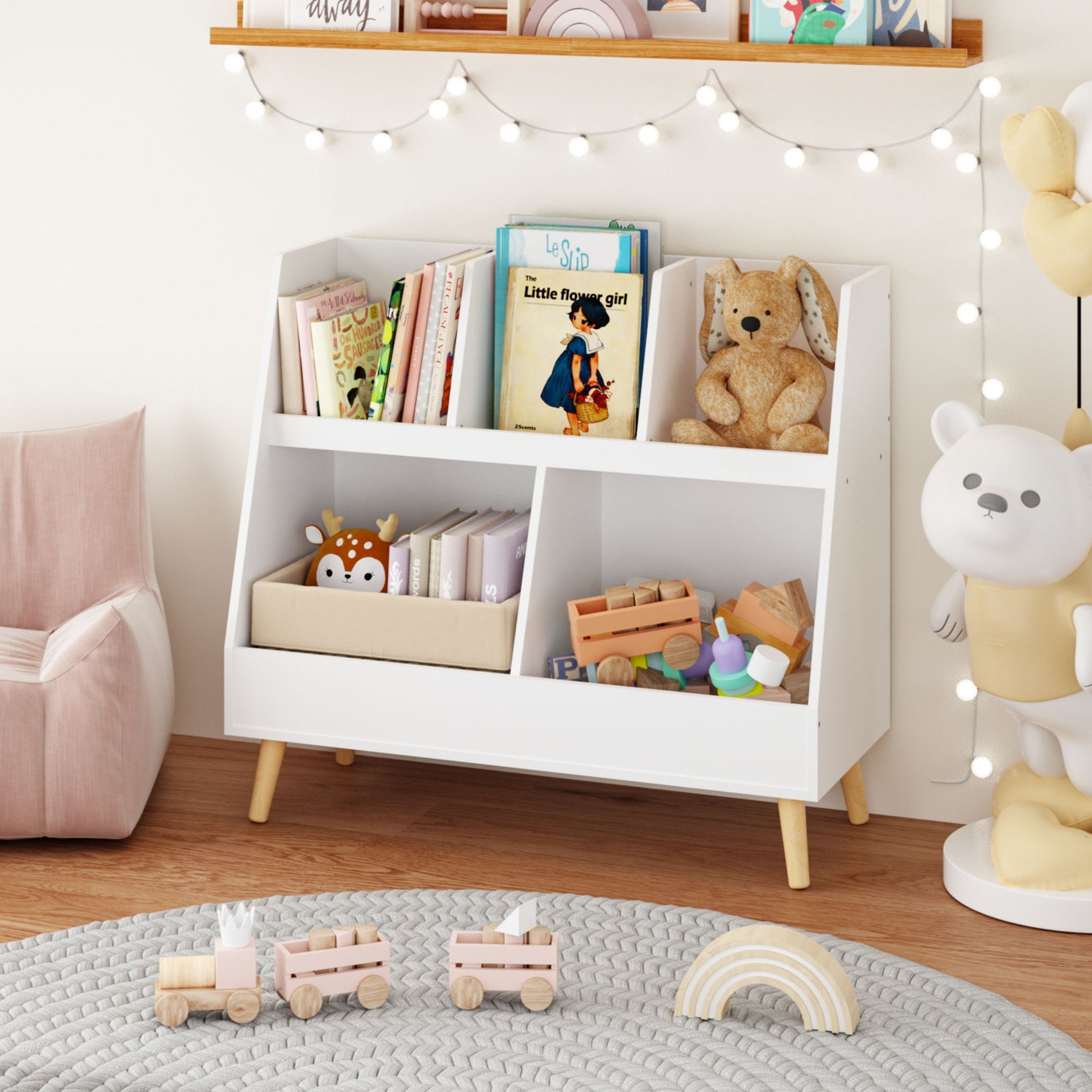 A children's playroom with a Kids Bookshelf And Toy Organizer for books, stuffed animals, and toys. To the left is a light pink chair. A gray rug with wooden train toys lies on the floor, and string lights hang above on the wall.