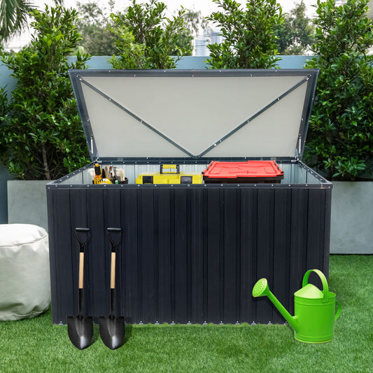 The Out Door Metal Storage Box Store Medium - Black sits on a grass lawn, containing various tools and a red container. Two shovels hang on its side, with a green watering can in front. Lush greenery and a white pouf are in the background.