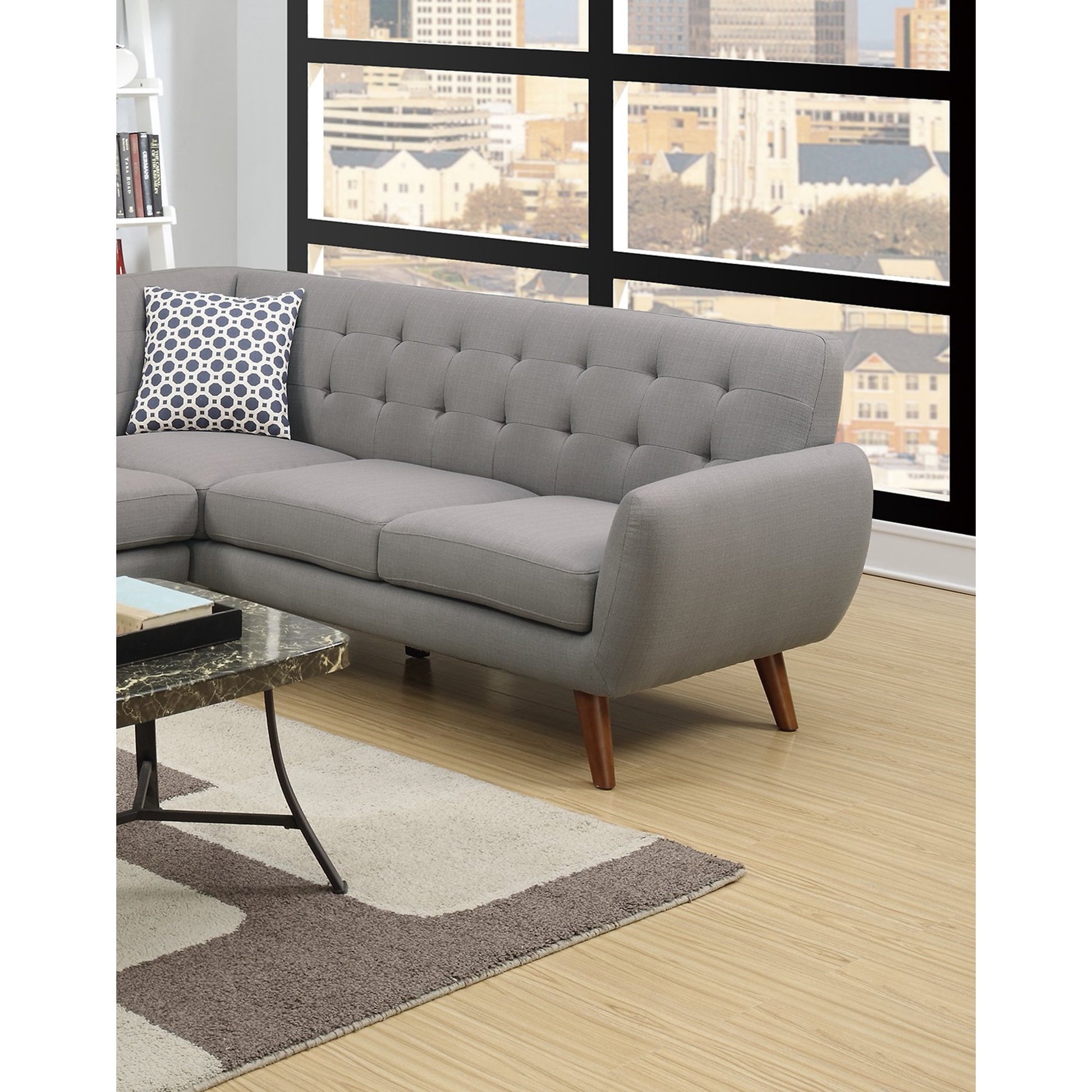 A modern living room features the Retro Modern Grey Sectional Sofa with wooden legs, placed next to a window revealing a cityscape view. On the sofa is a black and white polka dot pillow. In the foreground, there is a marble coffee table paired with a beige rug.