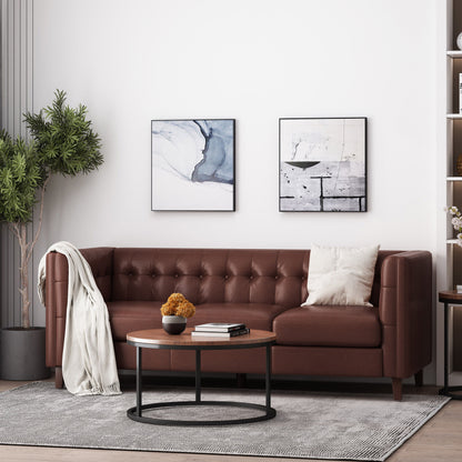 A modern living room features the Comfy 3 Seat Sofa with a brown leather finish, adorned with a white pillow and throw. Above it are two abstract paintings. A round wooden coffee table with a black frame and plant is centered, with a tall green plant on the left and shelves on the right.