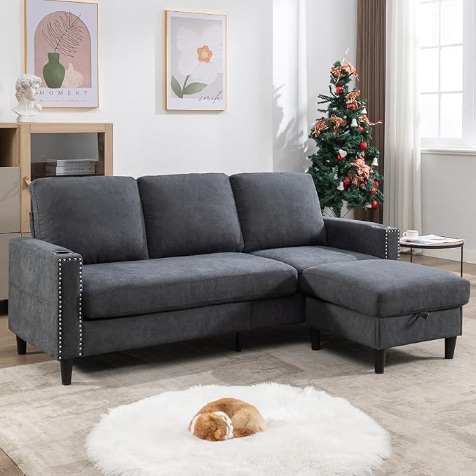 A cozy living room with the Siiejia Convertible Sectional L-Shaped Apartment in dark gray, adorned with studded accents. A small Christmas tree stands in the corner, and wall art hangs above the couch. A fluffy rug with a small dog curled up on it completes the scene.