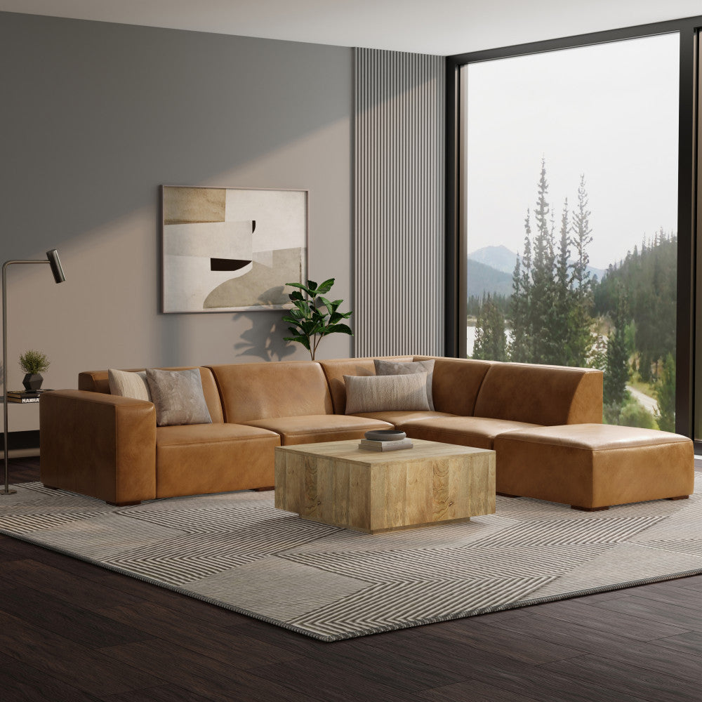 A modern living room with a large tan leather sectional sofa and a wooden coffee table. Gray and black patterned rug on dark wood floor. Large window with a view of trees and mountains. Minimalist artwork and a potted plant complete the decor.