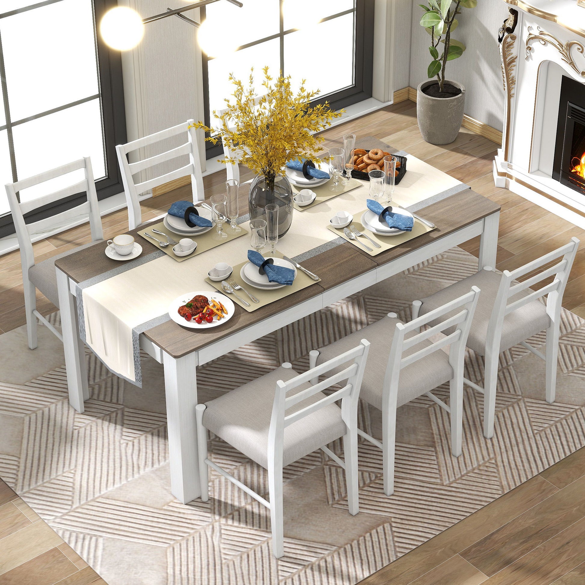 A dining room features an extendable wooden table with a cream runner, blue napkins, plates, cutlery, and a dark vase of yellow flowers. Six white cushioned chairs surround it. A patterned rug lies beneath near a fireplace.