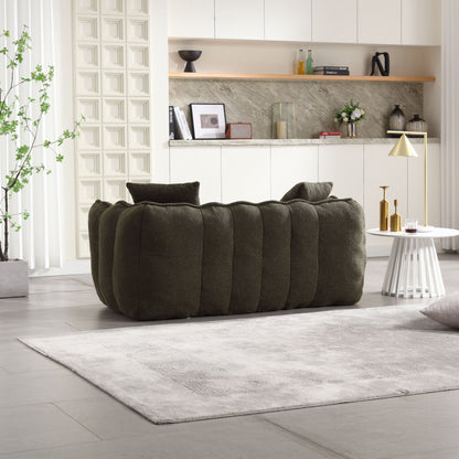 A modern living room features a dark green textured sofa facing away. A gray rug is on the floor, with a Soft Beanbag Chair for two. On a white side table sits a gold lamp, while decorative items adorn built-in shelves. A tall potted plant stands to the left.