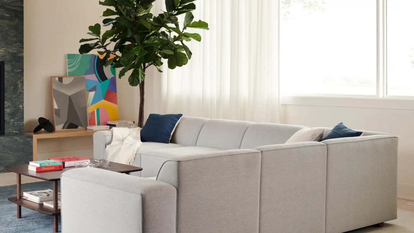 A modern living room featuring the Mambo Loveseat in light gray, complemented by blue pillows. A tall potted plant stands nearby, and a colorful abstract painting is partially visible on the wall. Bright natural light streams in through sheer curtains.