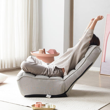 Relaxing on a Reclining Japanese Chair Lazy Sofa Tatami Balcony Adjustable Chair, a person stretches cross-legged in a bright room. They're dressed in light gray and next to them is a small round pink table. Sheer white curtains let sunlight stream through gracefully.