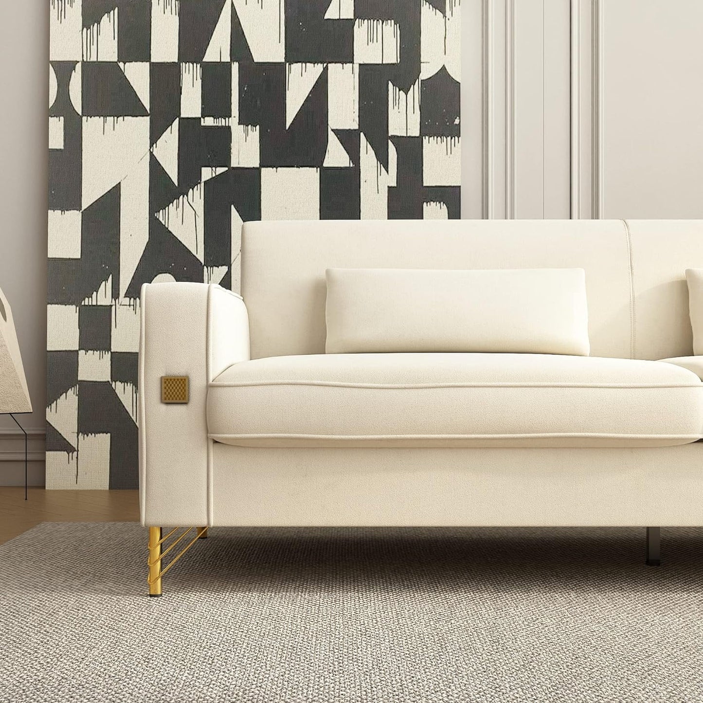 A modern living room showcases the Mid Century Modern Sofa Couch, which is cream-colored and features a rectangular cushion with sleek gold legs. The background displays a black and white geometric patterned wall, while a light gray rug covers the floor.