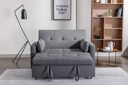 A modern gray 2-seater sleeper sofa bed with two cushions sits in a living room. A sleek floor lamp is to the left, and a wooden bookshelf stands on the right. A framed art print hangs above the sofa. Nearby, a round table holds a teapot and cup.