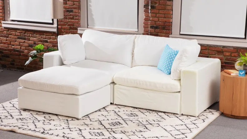 A cozy living room features a white sectional sofa with a chaise lounge, adorned with a blue polka-dotted accent pillow. The sofa sits on a patterned white rug, and the room has exposed brick walls and large windows with white blinds, allowing natural light in.
