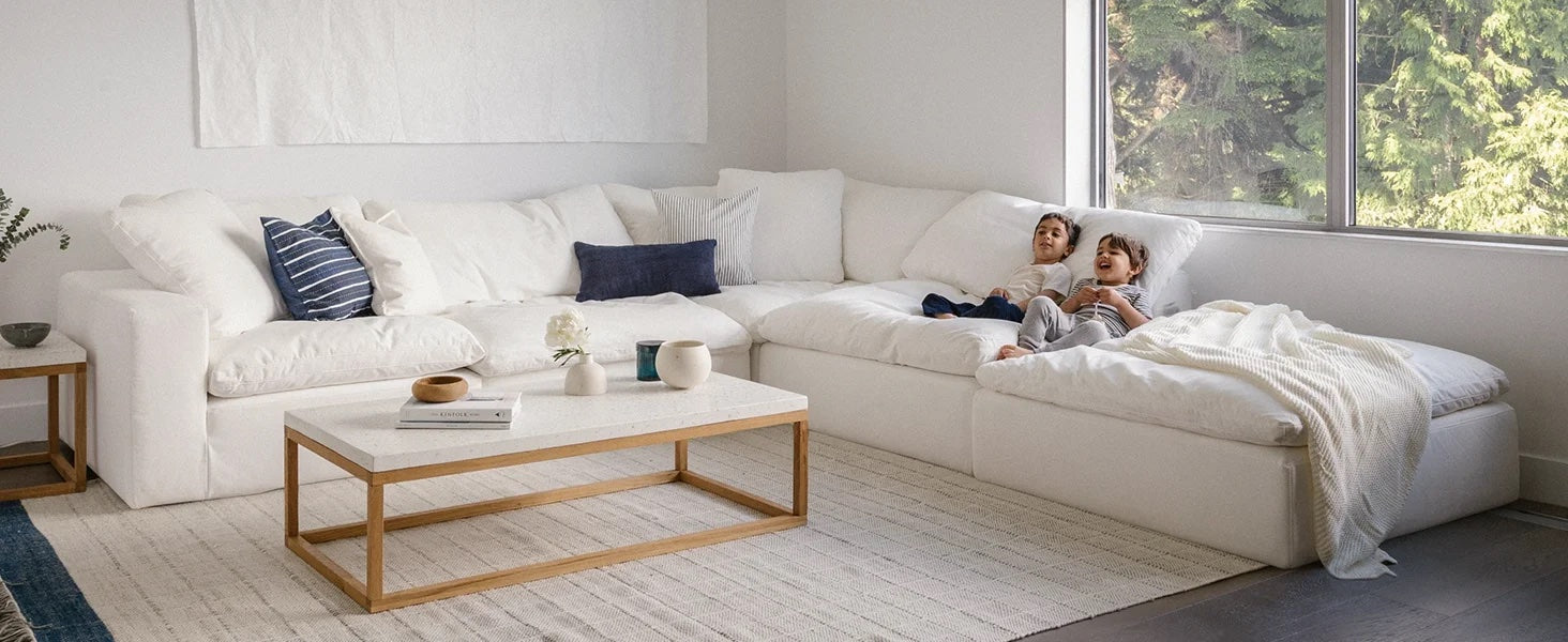 A cozy and spacious living room with a white sectional sofa, decorated with blue and white throw pillows. Two children sit together, relaxing. A light-colored coffee table with various decor items sits on a large area rug. A large window reveals greenery outside.
