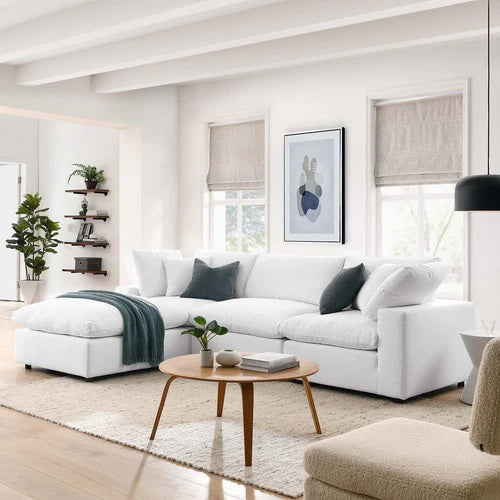 A modern living room with a white sectional sofa adorned with green and gray cushions. A green throw is draped over one arm. A wooden coffee table sits in the center, and a wall with two windows features artwork. There's a shelf with plants in the background.