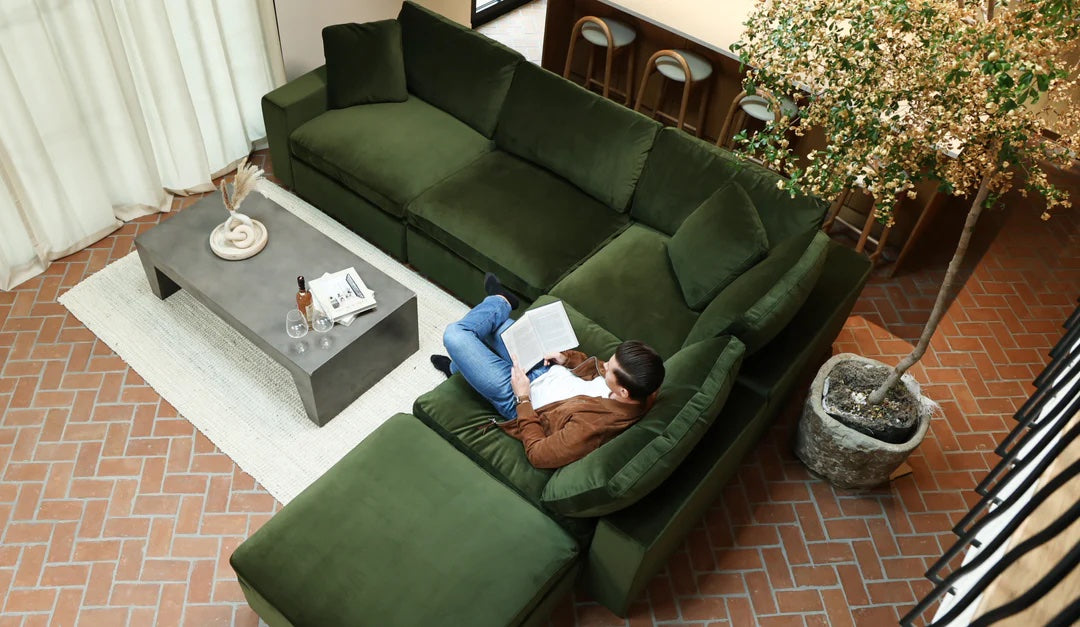 A person is lying on a large green sectional sofa, reading a book in a living room with brick flooring. Nearby is a concrete coffee table with a bottle, glasses, and decor. A tall indoor plant is placed beside the sofa, and barstools line the kitchen counter.