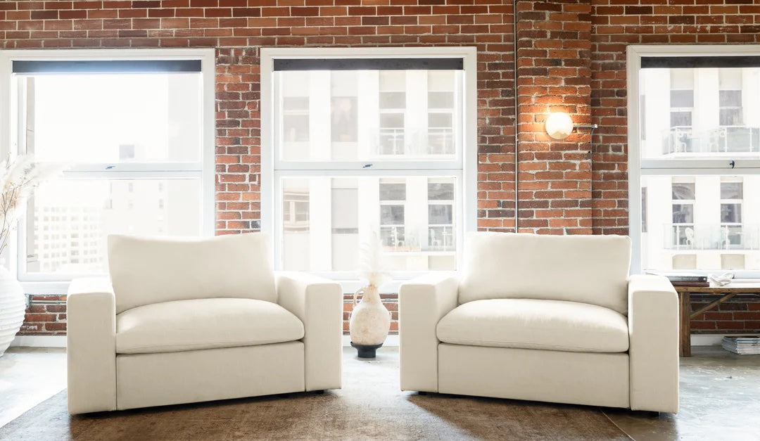 A stylish living room features two matching white armchairs positioned on a dark rug. The space is accented by three large windows with a view of city buildings, exposed brick walls, and a lit wall sconce. A small decorative item sits between the armchairs.