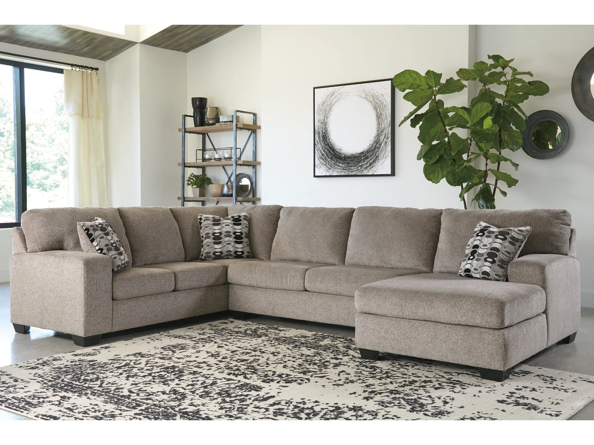 A spacious living room features a large beige Ballinasloe 3-Piece Sectional with Chaise decorated with patterned cushions. A modern black and white artwork hangs above, while a tall green plant and a wooden shelf sit in the background. The floor is covered by a patterned rug.