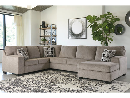 A spacious living room features a large beige Ballinasloe 3-Piece Sectional with Chaise decorated with patterned cushions. A modern black and white artwork hangs above, while a tall green plant and a wooden shelf sit in the background. The floor is covered by a patterned rug.