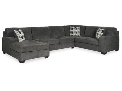 The Ballinasloe 3-Piece Sectional with Chaise features a gray L-shaped design with plush cushions and dark block legs. It includes two decorative pillows showcasing a geometric pattern, which add a touch of contrast. The chaise section is situated on the left side.