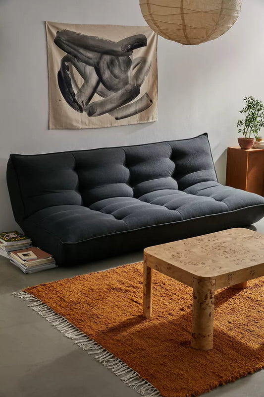 A cozy living room showcases the Greta XL Sleeper Sofa in dark gray, complemented by an abstract black and gray artwork on the wall. The space also includes a wooden coffee table, a rust-colored rug with fringe details, and a potted plant placed on top of a wooden cabinet. Above, a round paper lantern provides soft illumination.