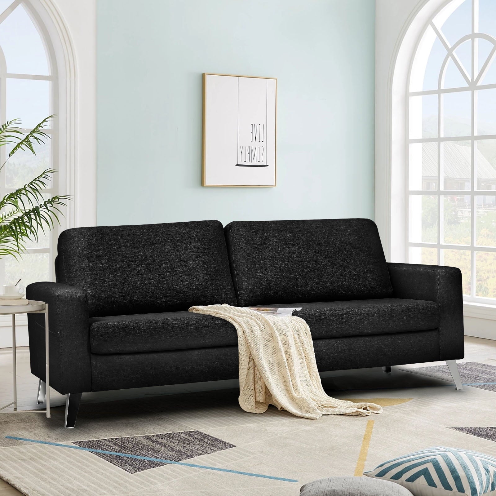 In a contemporary living room, the YODOLLA 79" Sectional Sofa in black is beautifully complemented by a light beige throw. The room features walls in a soft light blue shade and arched windows that invite natural light to fill the space. A plant positioned in the corner adds an element of greenery.