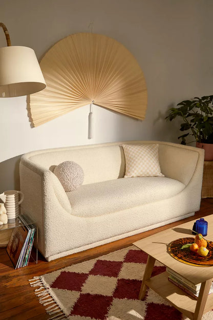 A cozy living room showcases an Aria Sofa in a cream color with two cushions, complemented by a large paper fan wall decoration. The space includes a wooden coffee table with a fruit plate and is illuminated by a floor lamp. In the corner, there is a plant, while part of the wooden floor is adorned with a red-and-white rug.